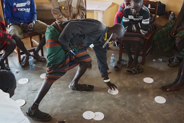 One participant stands in a room surrounded by chairs with other participants sitting and looking at him. On the floor are white paper circles with drawings. The standing participant is picking one.