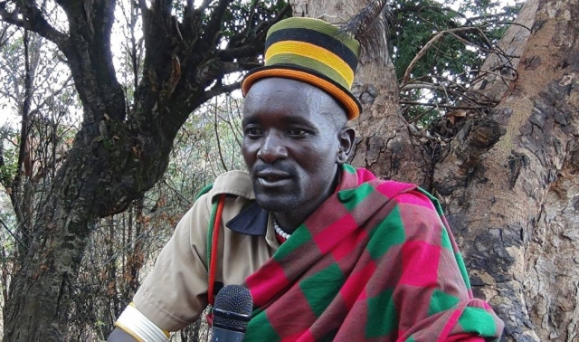 A portrait of Kamar, the vice chairman of Dodoth tribal group, holding a microphone, wearing a bright knitted hat with yellow and green colours as well as a tartan throw with green and pink checkers.