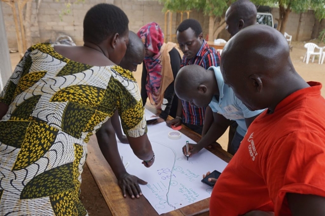 Several people gather around a table and one person is writing on a large flipchart. A storyline is visible with numbers and events. One woman wears a bright patterned African dress.