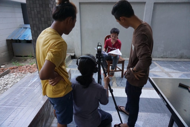 3 students stand around a tripod, filming a fellow student sitting on a chair.