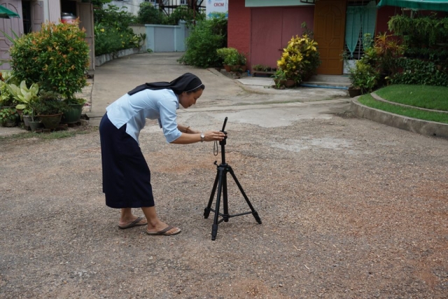 A woman in nun-attire is standing with a tripod filming in a courtyard