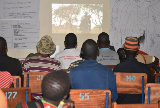 Back view of audience members watching a projection of films on a wall. Posters with drawings are hanging on either side of the projection. One t-shirt reads: Reconcile for development.